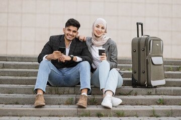 Wall Mural - Two multiracial business people sitting at stairs with travel bag and looking at camera. Diverse colleagues ready for the trip, sitting with phones and coffee on strairs outside