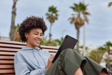 Wall Mural - positive inspired young woman with curly hair poses on wooden bench uses digital tablet and stylus m