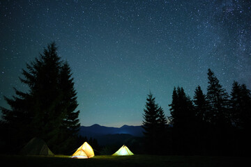 bright illuminated tourist tents glowing on camping site in dark mountains under night sky with spar