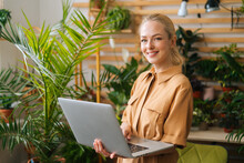 Portrait Of Cheerful Businesswoman Floral Store Owner Standing With Laptop Holding In Hand Among Green Plants, Looking At Camera. Pretty Female Florist Working Using Computer Of Greenhouse.