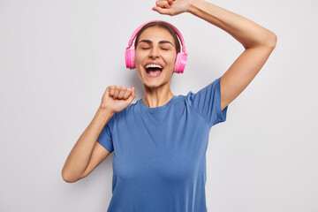 Wall Mural - Carefree brunette woman dances with rhythm of music wears stereo headphones on ears shakes arms smiles broadly dressed in casual blue t shirt isolated over white background. Positive meloman