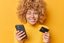 Positive Funny Curly Haired Woman Winks Eye Smiles Toothily Thinks Of Online Shopping Holds Mobile Phone And Banking Card Makes Payment Dressed In Casual Jumper Isolated Over Yellow Background.