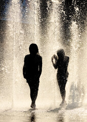 Silhouettes of a boy and a girl in the jets of cool fountain water on a hot summer day