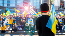 Young Boy Standing In Front Of A Crowd At A Protest Against The Russian Invasion In Ukraine. Child Holding On His Shoulder A Ukrainian Flag While Facing The Gathered People For A Peaceful Protest.