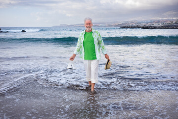 Happy senior attractive man standing barefoot on the shoreline at the beach holding shoes and hat in hands enjoying vacation Concept of relaxed and joyful retiree