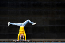 Athetic Young Woman Upside Down On A Skateboard, Black Wall Background, Yellow Shirt And Blue Jeans, Generation Z Female Crazy Lifestyle
