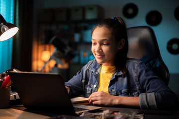 Focused smiling teen uses laptop at home, looking at screen, chatting, reading or writing email, paper for school