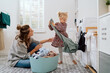 © ABCreative - Mom spends time with daughter in bathroom, laundry room while doing daily chores, girl takes things out of washing machine and hands to woman who puts colorful clean clothes into bowl