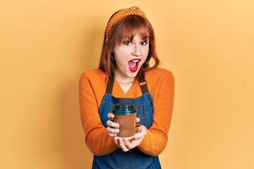 Poster - Redhead young woman wearing waitress apron holding take away cup of coffee celebrating crazy and amazed for success with open eyes screaming excited.