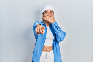 Wall Mural - Beautiful blonde woman wearing wool hat laughing at you, pointing finger to the camera with hand over mouth, shame expression