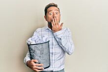 Middle Age Man Holding Paper Bin Full Of Crumpled Papers Covering Mouth With Hand, Shocked And Afraid For Mistake. Surprised Expression
