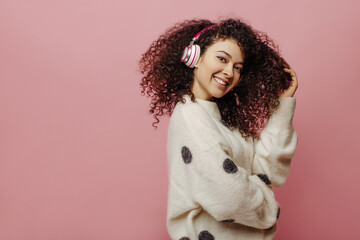 Beautiful caucasian young girl with curly hair listening to music with headphones on pink background in studio. Brunette smokes and wears warm white sweater. Beauty and youth concept