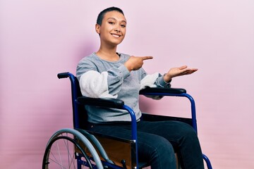 Sticker - Beautiful hispanic woman with short hair sitting on wheelchair amazed and smiling to the camera while presenting with hand and pointing with finger.