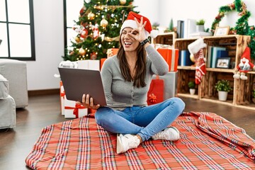 Sticker - Young latin woman using laptop sitting by christmas tree doing ok gesture with hand smiling, eye looking through fingers with happy face.