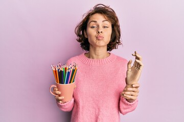 Sticker - Young brunette woman holding small wooden manikin hand and pencils puffing cheeks with funny face. mouth inflated with air, catching air.