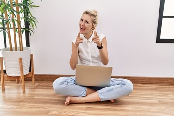 Poster - Young blonde woman using computer laptop sitting on the floor at the living room pointing fingers to camera with happy and funny face. good energy and vibes.