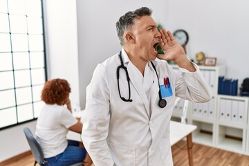 Wall Mural - Middle age doctor man at the clinic with a patient shouting and screaming loud to side with hand on mouth. communication concept.