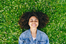 Smiling Afro Woman Lying On Green Flowering Plants On Meadow