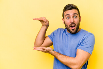 Young caucasian man isolated on yellow background shocked and amazed holding a copy space between hands.