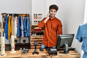 Wall Mural - Young hispanic shopkeeper man smiling happy talking on the smartphone working at clothing store.