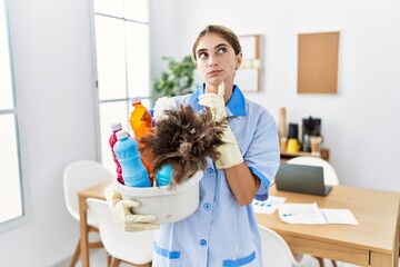 Canvas Print - Young blonde woman wearing cleaner uniform holding cleaning products thinking concentrated about doubt with finger on chin and looking up wondering