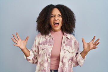 Wall Mural - Young african american woman standing over blue background crazy and mad shouting and yelling with aggressive expression and arms raised. frustration concept.