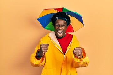 Poster - Young african american man wearing yellow raincoat angry and mad raising fists frustrated and furious while shouting with anger. rage and aggressive concept.