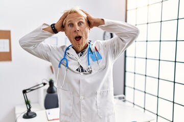 Poster - Young blond man wearing doctor uniform and stethoscope at clinic crazy and scared with hands on head, afraid and surprised of shock with open mouth