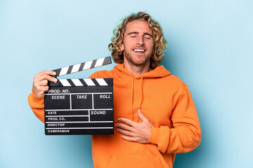 Wall Mural - Young caucasian man holding a clapperboard isolated on blue background laughs out loudly keeping hand on chest.