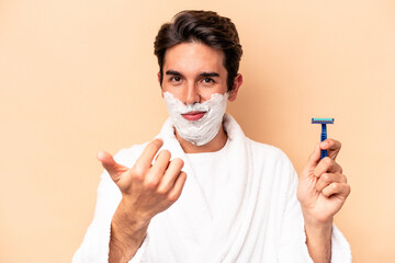 Wall Mural - Young caucasian man shaving his beard isolated on beige background pointing with finger at you as if inviting come closer.