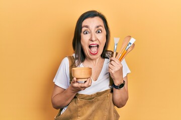 Wall Mural - Middle age hispanic woman holding makeup tools celebrating crazy and amazed for success with open eyes screaming excited.