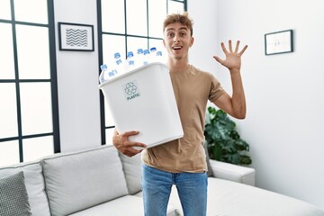 Wall Mural - Young caucasian man holding wastebasket with recycling plastic bottles at home celebrating victory with happy smile and winner expression with raised hands