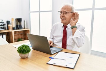 Wall Mural - Senior man working at the office using computer laptop smiling with hand over ear listening an hearing to rumor or gossip. deafness concept.