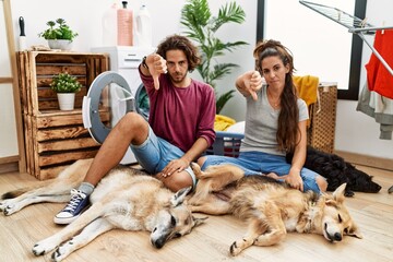 Sticker - Young hispanic couple doing laundry with dogs looking unhappy and angry showing rejection and negative with thumbs down gesture. bad expression.