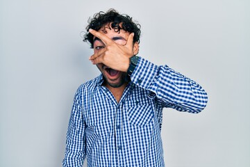 Sticker - Young hispanic man wearing casual clothes peeking in shock covering face and eyes with hand, looking through fingers with embarrassed expression.