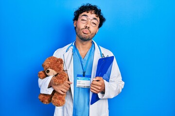 Poster - Young hispanic man wearing doctor uniform holding teddy bear and clipboard looking at the camera blowing a kiss being lovely and sexy. love expression.