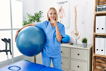 Sticker - Young caucasian physiotherapist woman holding pilates ball serious face thinking about question with hand on chin, thoughtful about confusing idea