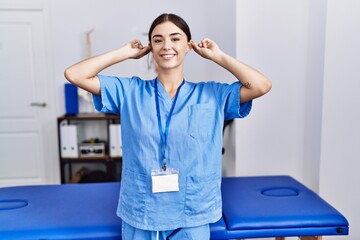 Poster - Young hispanic woman wearing physiotherapist uniform standing at clinic smiling pulling ears with fingers, funny gesture. audition problem