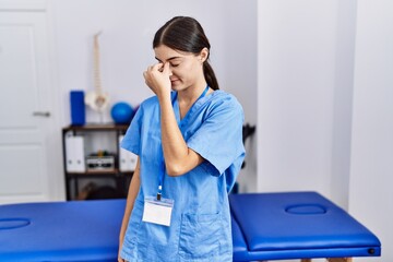 Sticker - Young hispanic woman wearing physiotherapist uniform standing at clinic tired rubbing nose and eyes feeling fatigue and headache. stress and frustration concept.