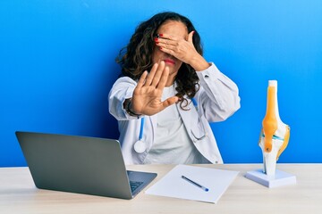 Canvas Print - Beautiful middle age woman doctor at orthopedic clinic covering eyes with hands and doing stop gesture with sad and fear expression. embarrassed and negative concept.