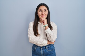 Sticker - Young latin woman standing over blue background with hand on chin thinking about question, pensive expression. smiling and thoughtful face. doubt concept.