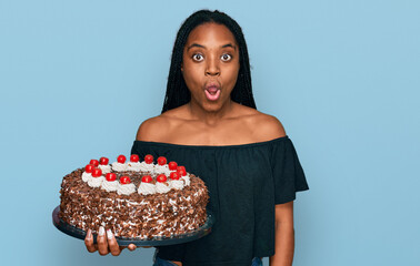 Wall Mural - Young african american woman celebrating birthday holding big chocolate cake scared and amazed with open mouth for surprise, disbelief face