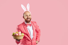 Studio Shot Of Happy Bald Young Man With Ginger Beard, Wearing Pink Suit, Bow Tie And Funny Bunny Ears Standing On Pink Background, Looking At Camera, Holding Wicker Bowl Of Easter Eggs And Laughing