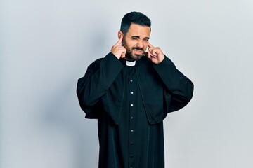 Poster - Handsome hispanic man with beard wearing catholic priest robe covering ears with fingers with annoyed expression for the noise of loud music. deaf concept.