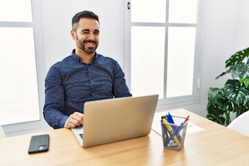 Sticker - Young hispanic man with beard working at the office with laptop winking looking at the camera with sexy expression, cheerful and happy face.