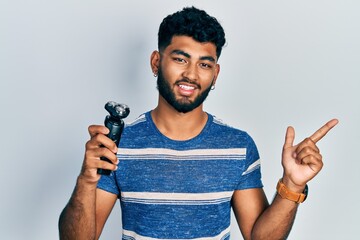 Wall Mural - Arab man with beard holding electric razor machine smiling happy pointing with hand and finger to the side