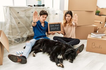 Canvas Print - Young caucasian couple with dog holding our first home blackboard at new house doing stop sing with palm of the hand. warning expression with negative and serious gesture on the face.