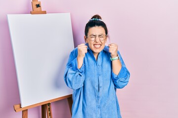 Poster - Young hispanic woman standing by painter easel stand excited for success with arms raised and eyes closed celebrating victory smiling. winner concept.