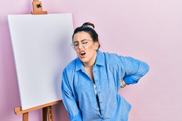 Poster - Young hispanic woman standing by painter easel stand suffering of backache, touching back with hand, muscular pain