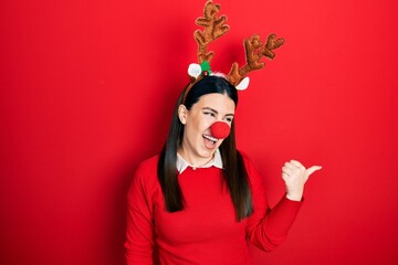 Canvas Print - Young hispanic woman wearing deer christmas hat and red nose smiling with happy face looking and pointing to the side with thumb up.